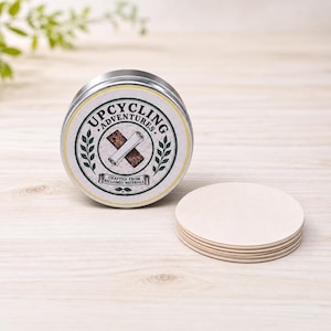 May include: A round silver metal tin with a label reading Upcycling Adventures Crafted From Reclaimed Materials, featuring a graphic of a scroll on wood. Beside the tin is a stack of several plain, circular, light-colored wooden discs.