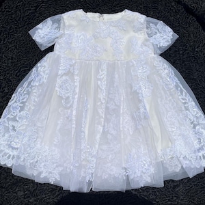 May include: A cream-colored dress featuring short sheer sleeves and a round neckline. The bodice and skirt are adorned with intricate white floral lace embroidery layered over a solid fabric base, creating a textured and layered appearance.