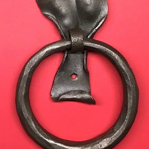 May include: A black metal door knocker shaped like a leaf with a ring handle. The knocker is mounted on a red background.