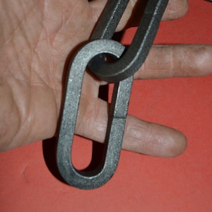 May include: A close-up of a black metal chain link. The chain is thick and heavy, and the links are interlocked. The chain is lying on a red surface.