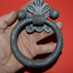 May include: A black metal ring door knocker with a decorative floral design and a textured ring. The knocker is held in a hand.