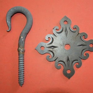 May include: A metal hook with a decorative, ornate,  square-shaped plate. The hook is attached to a threaded rod. The plate has a floral design and is made of metal.
