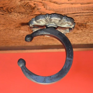May include: A decorative, dark metal hook with a floral design base, mounted on a wooden surface. The hook has a curved shape with rounded ends. The background is a solid red color.
