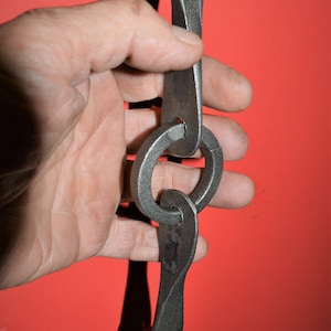 May include: A close-up of a hand holding a black metal chain with unique, hand-forged links. The chain is made of thick, sturdy metal and has a rustic, industrial look.