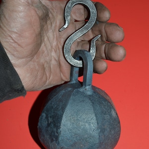 May include: A black metal weight with a rounded, faceted shape and a silver S-shaped hook. The weight is held in a hand.