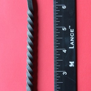 Peut inclure: Crochet en fer forgé à la main avec une section centrale torsadée et des extrémités recourbées. Le crochet mesure environ 20 cm de long et est présenté sur fond rouge avec une règle.
