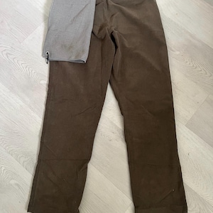 May include: Brown corduroy pants with a gray towel hanging from the back pocket. The pants have a classic, straight-leg design and a button and zipper closure. The towel is rectangular and has a grommet.