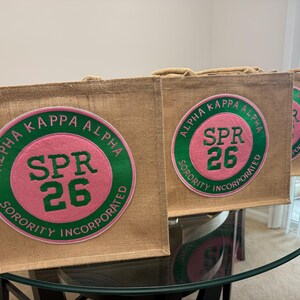 May include: Several tan burlap tote bags with a circular patch featuring the text "ALPHA KAPPA ALPHA SPR 26 SORORITY INCORPORATED" in green and pink. The bags are displayed on a glass table.