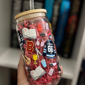 May include: A clear glass tumbler with a bamboo lid and a glass straw, filled with red paper shreds. The exterior features colorful decals of a hockey jersey with the text DI LAURENTIS 66, an open book, a gold trophy, a guitar, and headphones.