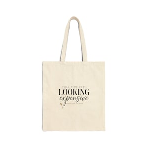 May include: Cream-colored tote bag with black text that reads "FULL-TIME JOB: LOOKING expensive" in a stylish font. The bag also has the text "fashion world, city edition" below the main text. The bag has two handles.