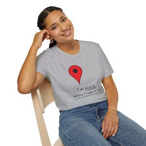 May include: A light gray t-shirt featuring a red location pin graphic with a black dot. Below the graphic, the text reads "I am exactly where I need to be." The shirt is made of a soft material.