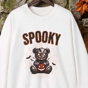 Puede incluir: Una sudadera blanca de manga larga con un estampado gráfico de un oso de peluche oscuro y amenazante que sostiene una pequeña calabaza. El oso está centrado en un diseño de telaraña, con la palabra SPOOKY impresa en letras grandes, negritas y de color marrón anaranjado.