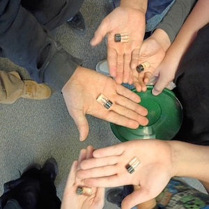 May include: Several small glass vials with black and gold caps are held in open hands. The vials appear to contain a clear liquid. The hands are of various sizes and skin tones. A green object is visible in the background.