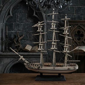 May include: A detailed model ship with three masts, painted in white and beige, sits on a dark wooden surface. The ship has intricate rigging and is displayed against a backdrop of a gothic-style fireplace and dark stone wall.