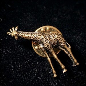 Puede incluir: Un pin de solapa dorado con forma de jirafa. El pin presenta un diseño detallado de jirafa con un patrón texturizado en su cuerpo y un respaldo de alfiler. La jirafa se representa de pie, con su largo cuello y patas.