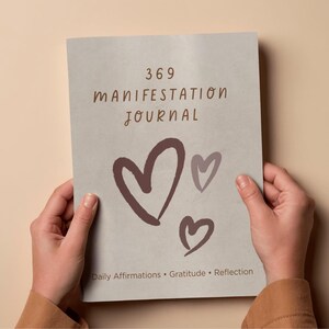 Peut inclure: Un journal avec le texte "369 Manifestation Journal" et trois illustrations de cœurs bruns. Le journal comprend également les mots "Daily Affirmations • Gratitude • Reflection". Le journal est tenu par deux mains.