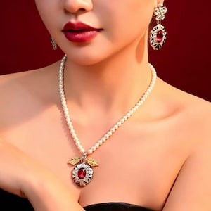 May include: A pearl necklace with a pendant featuring a large, oval, red gemstone. The pendant is accented with gold-coloured leaves and a silver-coloured frame. Matching earrings with red gemstones and floral designs complete the set.