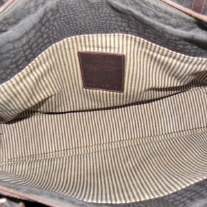 May include: The inside of a brown leather bag with a striped fabric lining. A brown leather tag with the word "Cole Haan" is visible.