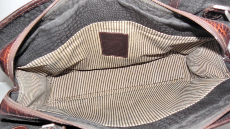 May include: The inside of a brown leather bag with a striped fabric lining. A brown leather tag with the word "Cole Haan" is visible.