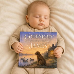 Op de afbeelding: Een slapende baby in een beige onesie houdt een kinderboek vast met de titel "Goodnight Little Duck Hunter JAMES". De boekomslag toont een tafereel aan het meer met een hond, eenden en een huis bij zonsondergang. De baby ligt op een crèmekleurige deken.