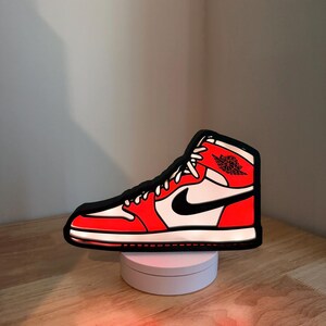 May include: A high-top sneaker-shaped LED light featuring a vibrant red, white, and black color scheme. The design includes a black Nike swoosh logo on the side and the Air Jordan wings emblem near the collar, all outlined in a bold black border.