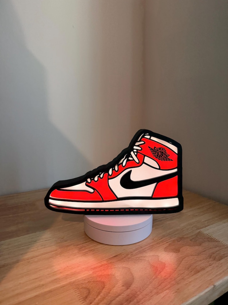 May include: A high-top sneaker-shaped LED light featuring a vibrant red, white, and black color scheme. The design includes a black Nike swoosh logo on the side and the Air Jordan wings emblem near the collar, all outlined in a bold black border.