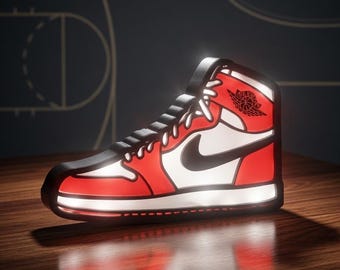 Puede incluir: Una lámpara iluminada con forma de zapatilla de deporte alta en rojo, blanco y negro. El diseño presenta un swoosh de Nike negro, cordones blancos y un logotipo de Air Jordan negro en el lateral. La lámpara emite un brillo desde el interior.