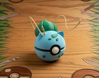 Puede incluir: Una Pokeball azul claro con temática de Bulbasaur con detalles verdes y un botón blanco. El diseño incluye una franja negra y puntos verdes. El fondo presenta diseños de Pokeball de madera, cuchillos y hierba verde.