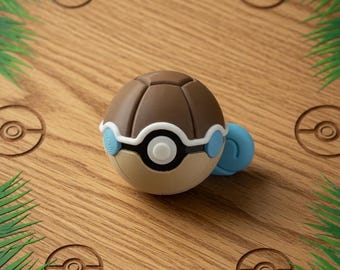 Puede incluir: Una Poké Ball marrón y beige con detalles azules, colocada sobre una superficie de madera con diseños de Poké Ball grabados y detalles de hojas verdes. La bola tiene una franja blanca y un centro negro.
