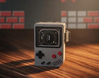 Puede incluir: Un reloj inteligente gris diseñado para parecerse a una consola de juegos portátil retro. Cuenta con una cruceta negra en forma de cruz, botones rojos y una pantalla que muestra "Ford Car Home". El reloj está sobre una superficie de madera.
