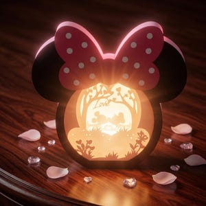 May include: A decorative lamp shaped like a mouse head with a pink polka-dot bow. The centre features a backlit cutout scene of two figures kissing under a tree with the word Love written in script. The lamp emits a warm glow from within.