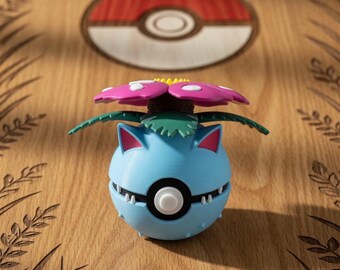 Puede incluir: Figura de Venusaur azul y blanco con una flor rosa y hojas verdes. La figura está sobre una superficie de madera con un diseño de hierba tallada y un logotipo de Pokeball en el fondo. La figura tiene un cuerpo redondo.