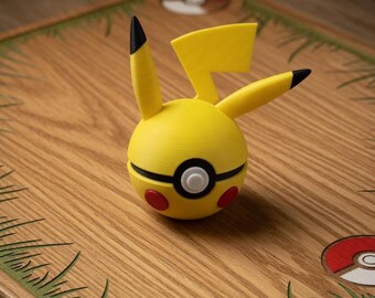 Puede incluir: Una figura amarilla de Pikachu con orejas y brazos con puntas negras, mejillas rojas y una raya negra. La figura está sobre una tabla de madera con hierba verde y diseños de Poké Ball rojos y blancos.