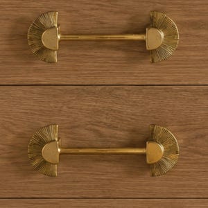 May include: Close-up of a wooden dresser with three drawers, each featuring a gold-colored, half-circle handle with a linear, textured design. The handles are centered on the drawers, providing a decorative and functional element.
