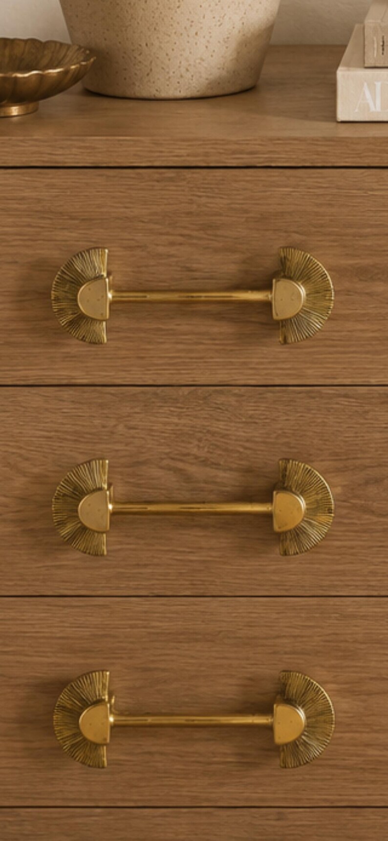 May include: Close-up of a wooden dresser with three drawers, each featuring a gold-colored, half-circle handle with a linear, textured design. The handles are centered on the drawers, providing a decorative and functional element.