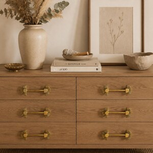 May include: A light brown wooden dresser with six drawers, each featuring gold-toned hardware. The dresser is adorned with decorative items, including a vase with dried flowers, a framed botanical print, and a small bowl.