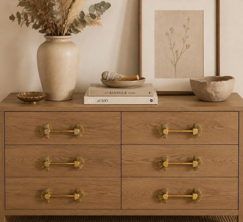 May include: A light brown wooden dresser with six drawers, each featuring gold-toned hardware. The dresser is adorned with decorative items, including a vase with dried flowers, a framed botanical print, and a small bowl.