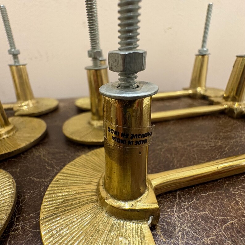 May include: Close-up of several brass-colored cabinet pulls with a semi-circular base and a cylindrical post. Each pull has a threaded bolt and nut for installation. The pulls are labeled "MADE IN INDIA".
