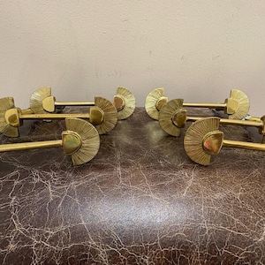 May include: A collection of six brass curtain rod holders. Each holder features a fan-shaped design with a central post and a long, straight rod. The holders are arranged on a textured, brown surface.