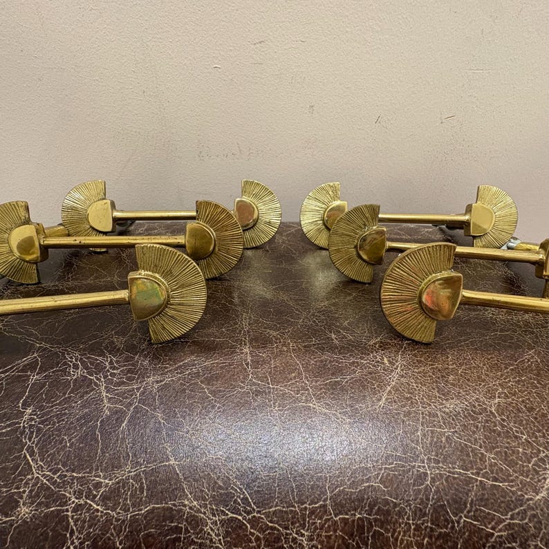 May include: A collection of six brass curtain rod holders. Each holder features a fan-shaped design with a central post and a long, straight rod. The holders are arranged on a textured, brown surface.