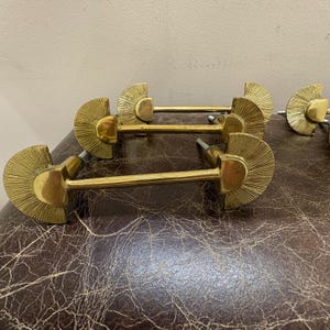 May include: A collection of brass cabinet pulls. Each pull features a fan-shaped design with a central handle. The pulls are arranged on a brown, textured surface, showcasing their vintage aesthetic and potential for furniture restoration.