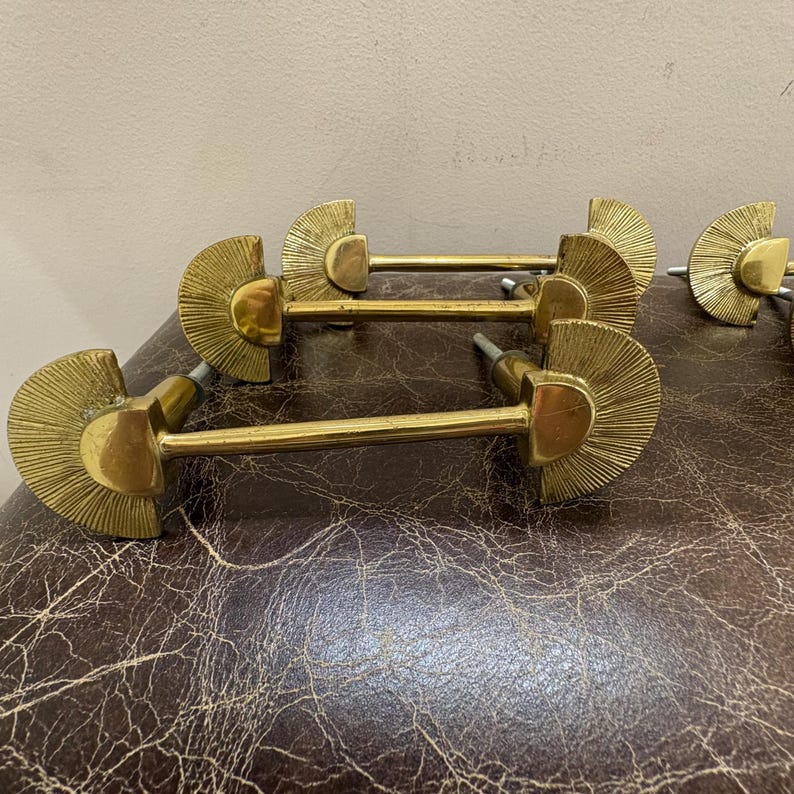 May include: A collection of brass cabinet pulls. Each pull features a fan-shaped design with a central handle. The pulls are arranged on a brown, textured surface, showcasing their vintage aesthetic and potential for furniture restoration.