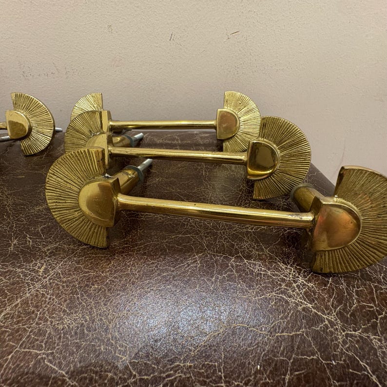 May include: A set of brass cabinet pulls. Each pull features a fan-shaped design with a central, rounded element and a long, straight handle. The pulls are a bright gold color and appear to be vintage.