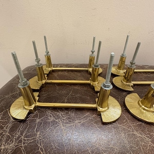 May include: A collection of brass-toned cabinet pulls. Each pull features a half-circle base with a ridged design, a cylindrical post, and a threaded screw for attachment. The pulls are arranged on a brown leather-like surface.
