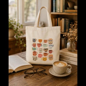 Puede incluir: Un bolso de tela color crema con un estampado gráfico de varias tazas coloridas de diferentes patrones y formas. El diseño incluye ilustraciones de tazas con motivos florales, de lunares y geométricos dispuestos en un patrón de cuadrícula.