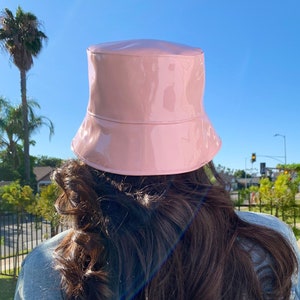 May include: A pale pink, shiny bucket hat. The hat has a glossy finish and a traditional bucket hat design. The hat is worn with a denim jacket. The background is a clear blue sky with a palm tree.