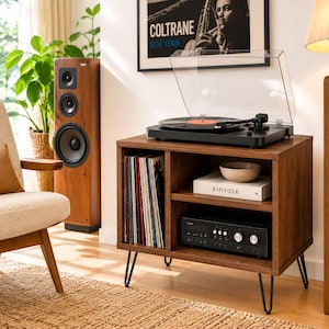 May include: A dark wood record player stand with black metal hairpin legs, featuring a vertical vinyl record storage compartment, an open shelf with a beige bowl and a Kinfolk book, and a lower shelf housing a black audio receiver.