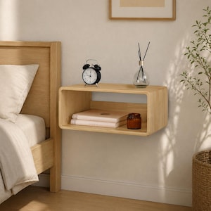 May include: A light-colored wooden floating shelf with rounded corners mounted on a wall. The shelf holds a black analog alarm clock, two stacked books with light-colored covers, and a small glass diffuser bottle with thin black reeds.