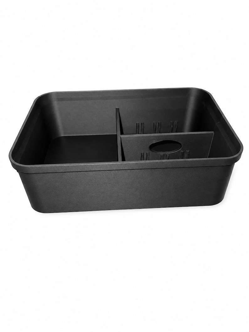 May include: A rectangular black plastic storage bin featuring a single internal divider that separates the interior into two distinct compartments. The divider includes a centered oval cutout and vertical ribbed detailing along its upper edge.