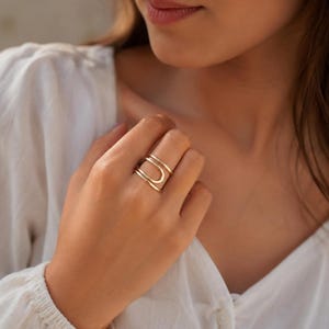 Op de afbeelding: Een goudkleurige ring met een dubbelbandig ontwerp en een gladde, gepolijste afwerking. De ring bestaat uit twee parallelle banden die buigen in een afgeronde, open vorm, waardoor een gelaagd uiterlijk op de vinger ontstaat met een strakke uitstraling.